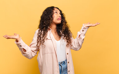 young hispanic woman shrugging with a dumb, crazy, confused, puzzled expression, feeling annoyed...