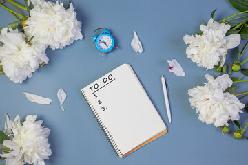 open notebook for writing plans, to-do list or dreams with fresh peonies, alarm clock, on light blue background.