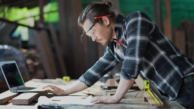 young hipster carpenter man working with computer laptop in workshop . craftsman wearing safety glasses checking order of clients or learning online