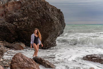 A beautiful girl in a black dress is walking on the waves, big waves with white foam. A cloudy stormy day at sea, with clouds and big waves hitting the rocks.