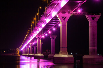Side view of colourful bridge illuminated with pink color lights at the night. Bridge stands on Volga river in Russia. Pink light is reflected in the water.