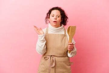 Woman with Down syndrome cooking at home isolated on pink background surprised and shocked.