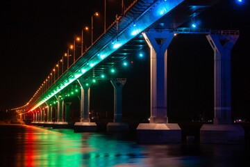 Low angle view of colourful bridge illuminated with different color lights at the night. Bridge stands on Volga river in Russia. Multi-colored light is reflected in the water.