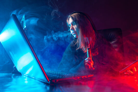 Girl is sitting at her laptop and smoking a vape. A teenager with a vape is filming a podcast. Girl blogger communicates with subscribers. A girl with electronic gadgets on a dark smoky background.