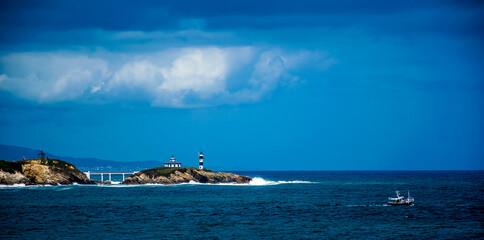 Un faro en una pequeña isla unida a tierra mediante un puente, un barco pesquero de color negro, nubes de tormenta y el mar cantábrico en la costa española