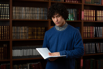 portrait of a young student boy in a blue sweater, standing with a book in his hands in the library against the background of books. Photo for advertising of university, college, student life.