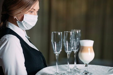 A female Waiter of European appearance in a medical mask serves Latte coffee.