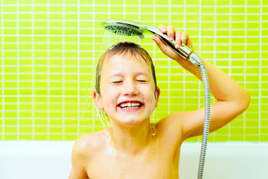 Boy in the bathtub washes his head with water sprayed from the shower head, with funny expression.