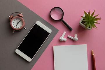 Modern workplace with smartphone, note pad, pen, alarm clock, plant, magnifying glass, and headphones on pink and grey geometric background. Business and education concept. Top view, flat lay