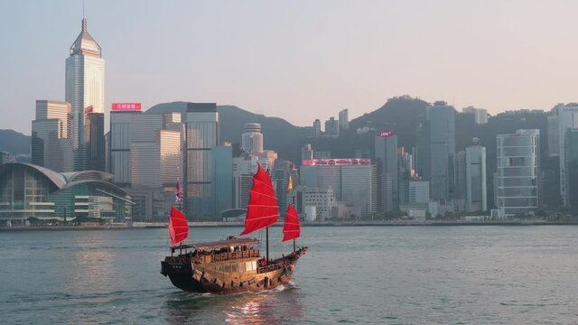 View of Victoria Harbour waterfront as a wooden red-sailed junk boat based on ancient Chinese sailing ships, now used as a touristic attraction, sails in front of the Hong Kong Island skyline.