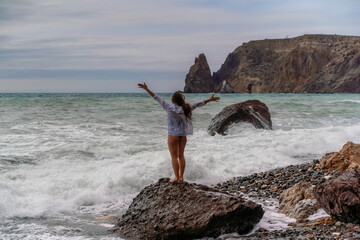 A beautiful girl in a white shirt and black swimsuit stands on a rock, big waves with white foam. A cloudy stormy day at sea, with clouds and big waves hitting the rocks.