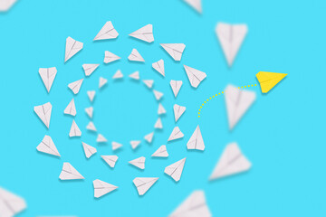 A yellow paper airplane flies out of the circle of white planes. Blue background. Flat lay. The concept of creativity and independent