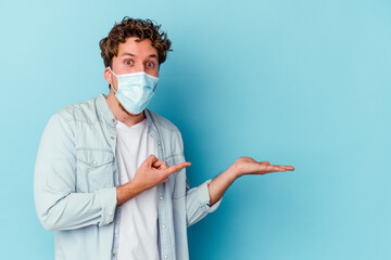 Young caucasian man wearing an antiviral mask isolated on blue background excited holding a copy space on palm.