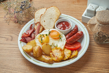 An appetizing breakfast option - fried egg with sausages, bacon and sausages, potatoes and corn, served in a white plate with red sauce. Wood background. Restaurant food