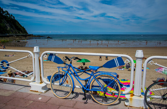 Una bicicleta aparcada junto a una valla azul con remos en una aldea costera del Cant&aacute;brico espa&ntilde;ol, con personas en la playa