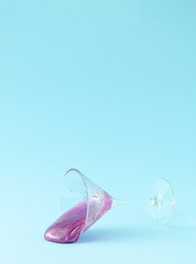 Purple glitter slime spilled out from crystal champagne glass on pastel blue background. Minimal party composition.