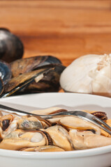 Top, close distance of a white bowl of freshly picked, local, steamed mussels, with a picking fork with shells off to a side and a bowl of clam juice and a garlic head, on a wood serving board