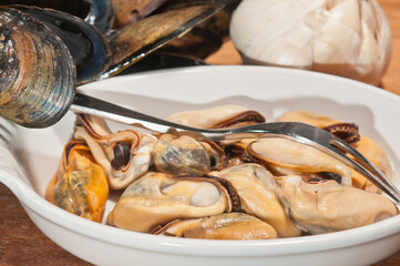 Top, close distance of a white bowl of freshly picked, local, steamed mussels, with a picking fork with shells off to a side and a bowl of clam juice and a garlic head, on a wood serving board