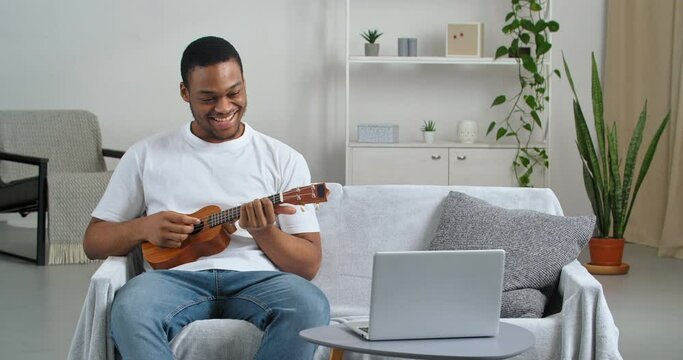 Afro american guy student wears casual clothes sitting on sofa with musical instrument playing ukulele composing melody making progress expresses joy with victory hand gesture, lock down homeschooling