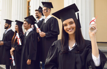 Young smiling girl university graduate standing holding diploma in raised hand over group of mates and university building at background. Graduation from university, education, diploma concept