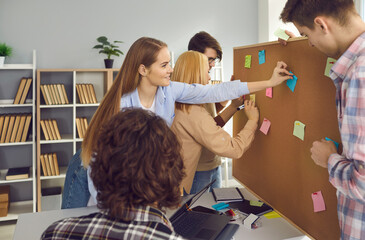 College or university students working on group project. Creative schoolmates brainstorming in school library, exchanging ideas, writing them on colorful sticky notes and pinning to bulletin board