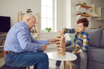 Senior aged mature man grandfather sitting and playing Jenga board game with his grandson boy at home with room interior at background. Grandchildren spending time with grandparents concept