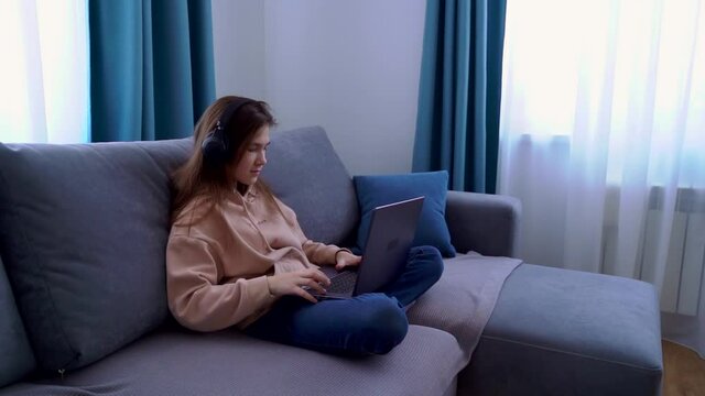 Smiling young girl in headphones using laptop while sitting on sofa at home. Happy girl looking at computer screen watching webinar, online language learning, distance education concept.