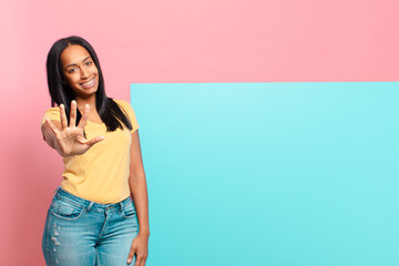 young black woman smiling and looking friendly, showing number five or fifth with hand forward,...