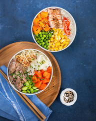 Two assorted poke bowls, flamed salmon, pulled pork, vegetables, rice, sauces. Top view, close-up. Hawaiian dish, blue dark background. Trendy asian food. Healthy and clean eating concept.