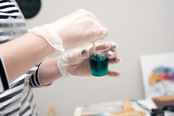 artist's hands in disposable transparent gloves, stirring a green dye in a plastic cup with epoxy...