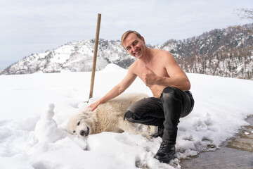 A man with a naked torso caresses a white dog in the snow