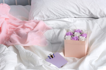 Box with white and violet roses lie on white cotton bed linen