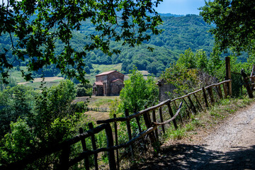 Fototapeta na wymiar Una iglesia prerrománica se asoma en un camino en las montañas cubiertas de bosques y prados con vallas de madera en la región española de Asturias