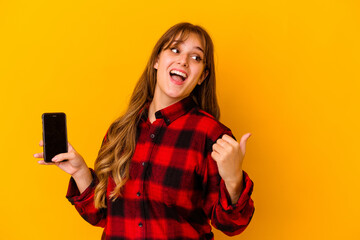 Young caucasian woman holding phone isolated on yellow background points with thumb finger away, laughing and carefree.