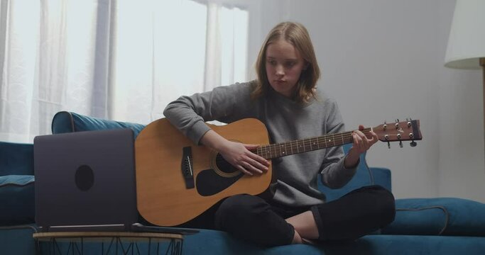 A young girl is sitting on the sofa in the lotus position in the living room. A woman learns to play the guitar alone. She is watching her first online lesson on her laptop.