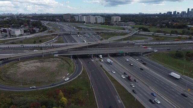 2.7K great aerial shot flying perpendicularly over 16 lanes of highway 401 traffic while seeing 2 subways go by each other in opposite directions and buildings in the background on horizon