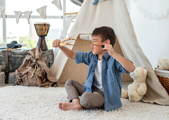 Little boy with wood slingshot sitting on room's floor in front of wigwam © tan4ikk