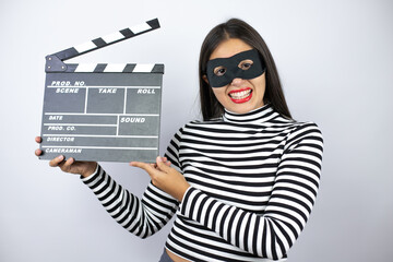 Young beautiful brunette burglar woman wearing mask holding clapperboard very happy having fun