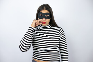 Young beautiful brunette burglar woman wearing mask mouth and lips shut as zip with fingers. Secret...
