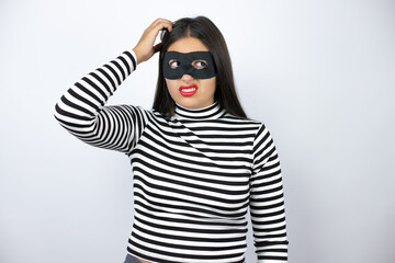 Young beautiful brunette burglar woman wearing mask confuse and wonder about question. Uncertain with doubt, thinking with hand on head