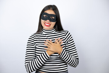 Young beautiful brunette burglar woman wearing mask smiling with her hands on her chest and...