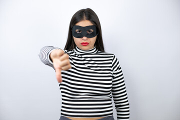 Young beautiful brunette burglar woman wearing mask with angry face, negative sign showing dislike...