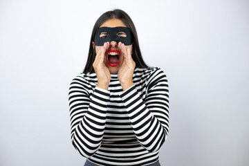 Young beautiful brunette burglar woman wearing mask shouting and screaming loud to side with hands on mouth