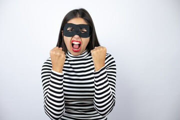 Young beautiful brunette burglar woman wearing mask very happy and excited making winner gesture with raised arms, smiling and screaming for success.