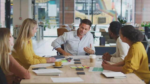 Businessman in wheelchair giving presentation on performance to colleagues sitting around table in modern open plan office - shot in slow motion