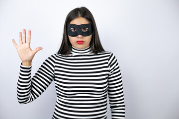 Young beautiful brunette burglar woman wearing mask showing and pointing up with fingers number five smiling confident and happy