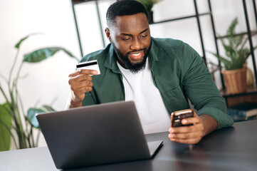 Happy smiling stylish african american guy sits at his desk at home or office, uses her smartphone...