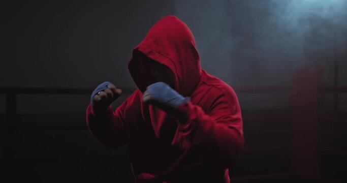 Portrait, a boxer in a hood is practicing blows, his hands are tied with protective bandages. Shadowboxing, dark ring with large tubers of smoke, close-up. 4k, ProRes