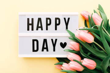 Happy Day words on lightbox with bouquet of pink tulips on light yellow background.