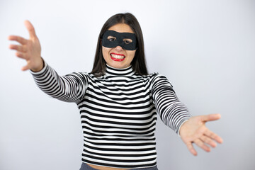 Young beautiful brunette burglar woman wearing mask looking at the camera smiling with open arms for hug